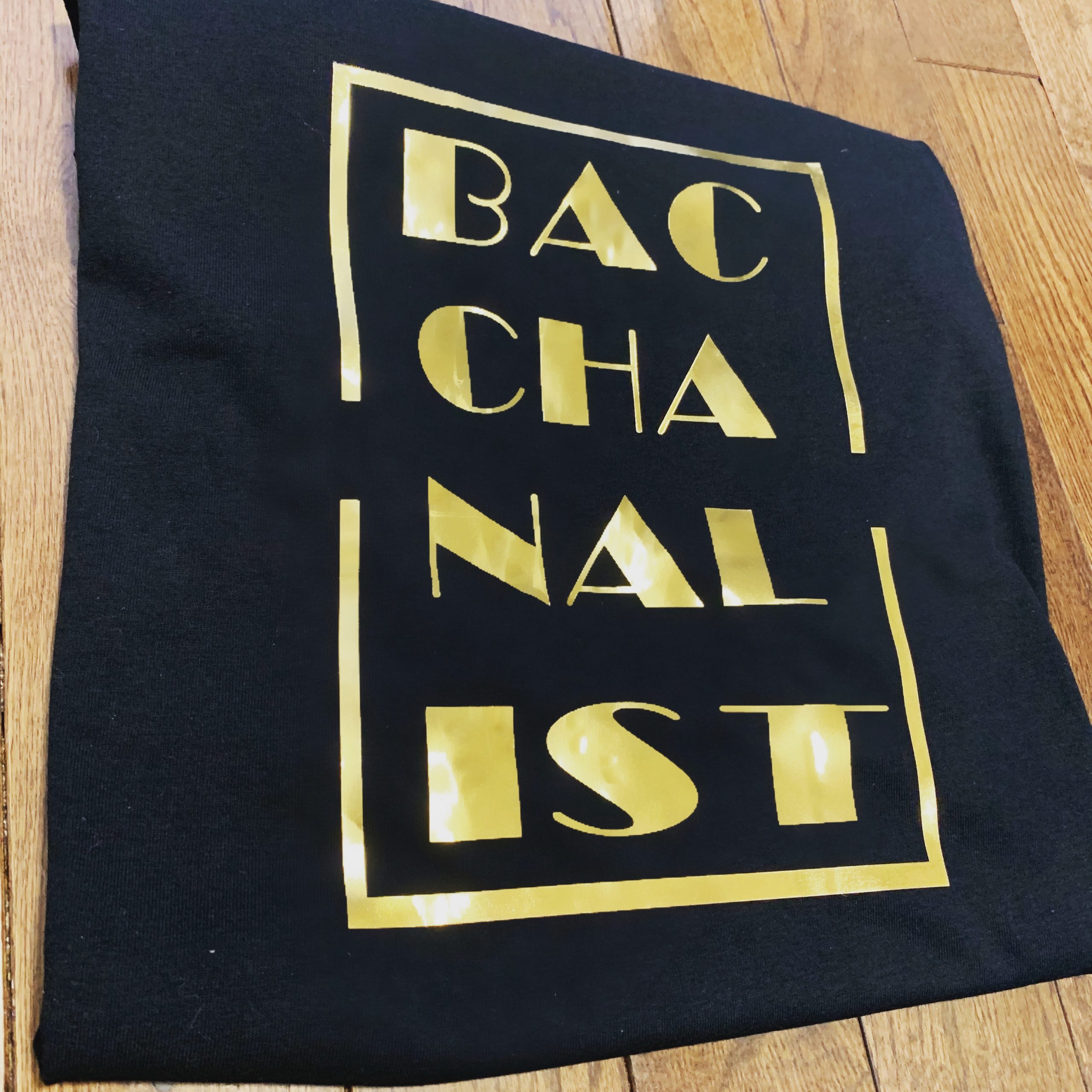 Black shirt with gold text that reads bacchanalist vertically divided into 4 sections of 3 letters at a time.